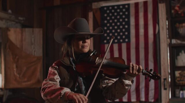 Woman Wearing Cowboy Hat and Vest Playing Fiddle Violin Music In Front of American Flag Inside of Old Rustic Saloon