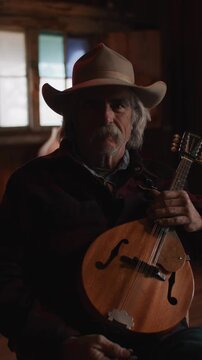 Man Wearing Cowboy Hat and Flannel Shirt Holding Mandolin String Instrument and Inside of Old Rustic Saloon