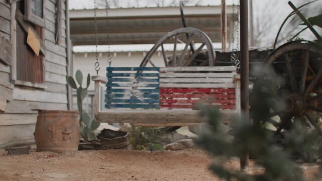 Focus On Texas Flag Painted Porch Swing Near Old Rustic Wagon Wheel and Cactus Outside of Old Saloon