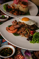 Close-up of Malaysian Char Kway Teow served with large prawns and sunny side up egg on a white plate, styled with chili sauce and traditional batik setting.