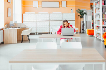 A young Caucasian woman with brown hair works on a laptop in a modern coworking office. The space features bright orange walls and wooden tables.