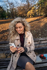 Beautiful curly hair girl using smartphone in the park on the bench, beautiful fall day