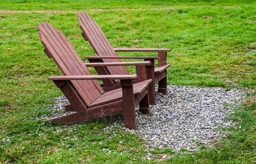 Two wooden Adirondack chairs on a small gravel area in a green grassy field, creating a peaceful outdoor seating background with copy space and nobody around.