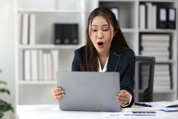 Young Asian businesswoman is working in her office and looking at her laptop with a shocked expression, surprised by unexpected news or data.