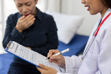 Female doctor wearing lab coat and stethoscope is writing down symptoms of a sick female patient coughing and sore throat, covering her mouth with her hand, during medical examination in hospital room