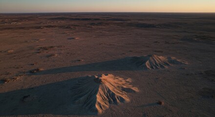 Aerial view of two eroded, sand-like mounds in a vast, desolate landscape at sunset