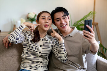 Asian young adult couple taking happy funny selfie smartphone social media casual home living room relaxing playful joyful digital lifestyle content sharing together
