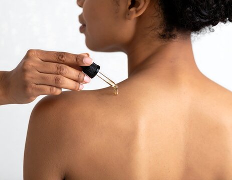 A person's bare back and shoulder receive skincare drops from a dropper held by another person's hand. Focus is on self-care
