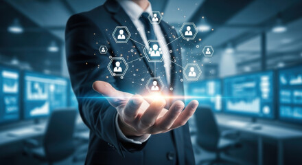 A businessman in a suit holds a glowing digital network in his open hand. Modern office with multiple monitors in the background.