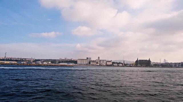 A scenic seascape of Istanbul's historic Sarayburnu point. A ferry crosses the water with the ancient walls of Topkapi Palace and lush trees in the background.