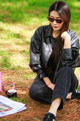 Young woman sitting on the grass in a park wearing sunglasses and a black leather jacket