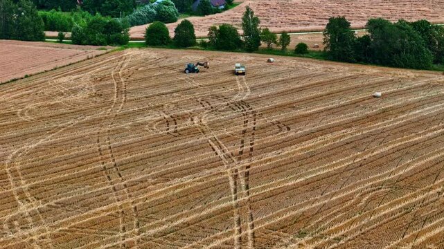 Tractor with front loader lifting hay bales onto trailer in harvested crop field