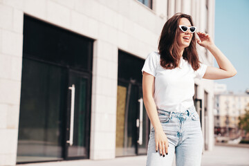 Fototapeta premium Young beautiful smiling hipster woman in trendy summer white t-shirt and jeans clothes. Sexy carefree model posing on the street background at sunset. Positive model outdoors, in sunglasses