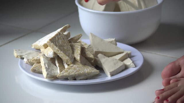 A close-up shot of hands arranging freshly cut tofu and triangular tempeh slices on a white plate, ready for seasoning and cooking. Tofu and tempeh are essential, high-protein staples in Indonesian.