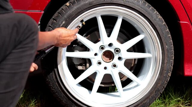 A close-up shot of a person using a tire inflator and pressure gauge on the valve stem of a red car's custom white and silver alloy wheel to check or add air pressure.
