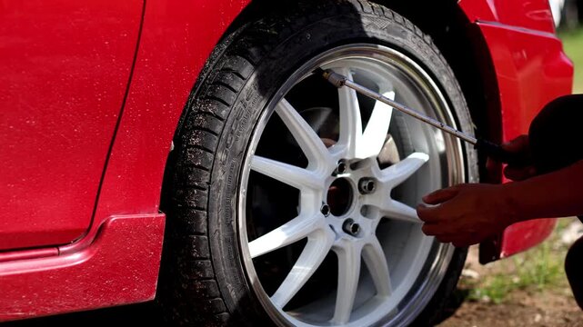 A close-up shot of a person using a tire inflator and pressure gauge on the valve stem of a red car's custom white and silver alloy wheel to check or add air pressure.
