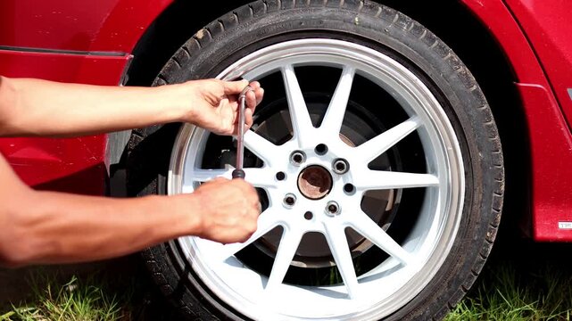 A close-up shot of a person using a tire inflator and pressure gauge on the valve stem of a red car's custom white and silver alloy wheel to check or add air pressure.