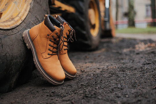 Pair of work boots leaning against a large construction vehicle wheel on a dirt ground. Durability, safety, and rugged industrial footwear.