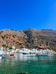 Obraz premium Loutro coastal village in Crete island Greece. White houses, moored boats on calm water below rocky hills, blue summer sky