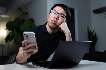 An Asian man wearing glasses holds his smartphone while sitting at his desk with a laptop. He appears thoughtful or bored, resting his head on his hand in a modern home office setting.