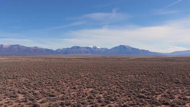 Aerial view moving across the arid sagebrush plains of Northern New Mexico toward the majestic Sangre de Cristo Mountains. Captures the distinct high desert terrain.