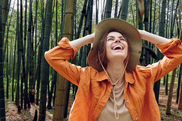 Woman wearing a hat laughing in a bamboo forest, outdoor nature portrait with joyful smile, casual orange jacket and relaxed carefree travel vibe captured in sunlight.