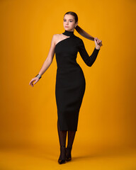 A full-length portrait of a young woman in an elegant, form-fitting black midi dress. She stands confidently, adjusting her high ponytail, against an orange background.