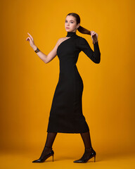 A full-length portrait of a young woman in an elegant, form-fitting black midi dress. She stands confidently, adjusting her high ponytail, against an orange background.