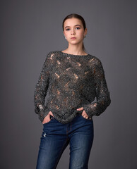 Young woman in a trendy shiny, open-knit sweater and jeans posing against a gray background. Casual glamour style.