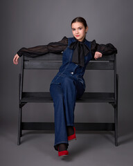 Studio fashion portrait of a young woman wearing a dark denim overalls or a set consisting of a structured vest and flared trousers, paired with a black sheer blouse, a tie and red high heels.
