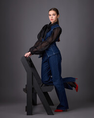 Studio fashion portrait of a young woman wearing a dark denim overalls or a set consisting of a structured vest and flared trousers, paired with a black sheer blouse, a tie and red high heels.
