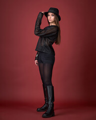 Dynamic studio fashion shot of a woman in a sheer black top, shorts, high boots, and fedora, walking against a vivid red background. Edgy, bold, and contemporary style.
