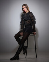 A studio fashion portrait of a young woman seated on a high chair, wearing a sheer black blouse with puffy sleeves, black shorts, tights, and chunky leather thigh-high boots. Against a light gray back