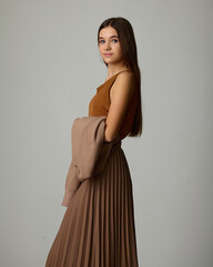 A close-up studio fashion portrait of a young woman wearing a monochrome brown ensemble consisting of a pleated midi skirt and a knitted cardigan draped over her arms. She poses with a calm expression