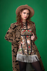 A portrait of a young woman in a brown velvet coat with a floral print and a mini-dress with fringe and a folk pattern, complemented by a fedora hat and beads.
