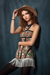 A fashionable portrait of a young woman in a stylish, modern bohemian outfit, wearing a brown fedora hat, posing against a dark, draped studio background.