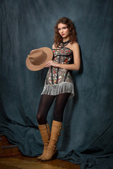 A studio portrait of a young woman standing full-length, wearing a brown fedora hat. She is wearing a strapless dress with a floral pattern, black tights, and tall brown boots.
