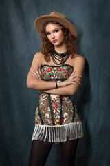 Close-up of a fashionable portrait of a young woman in a strapless dress with a bright floral print in a folk style. She is wearing a brown fedora hat and a black beaded necklace.