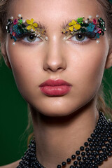 A striking close-up portrait of a young woman, adorned with intricate, colorful floral and pearl elements around her eyes. The high-fashion, fantasy makeup creates a vibrant, artistic image.