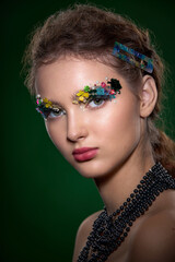 A striking close-up portrait of a young woman, adorned with intricate, colorful floral and pearl elements around her eyes. The high-fashion, fantasy makeup creates a vibrant, artistic image.