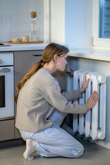 Upset freezing woman warming hands on heater at home during cold winter day. Sad female puts arms on radiator heating to warm up in kitchen. Heated season, coldness indoor, checks radiator temperature