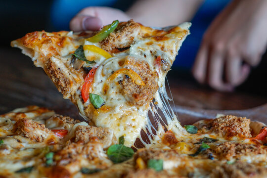 Close-up of a hand lifting a crispy chicken pizza slice with melted cheese pull, peppers, onions, and basil. Ideal for food ads, menus, delivery promotions, and culinary marketing