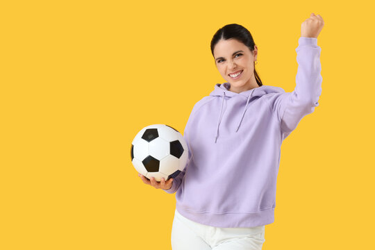 Happy young woman with soccer ball celebrating success on yellow background. Sports bet concept