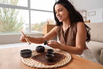Beautiful young woman putting dry tea into teapot at table in room
