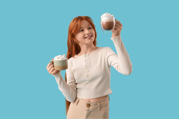 Teenage redhead girl with cups of sweet cocoa on blue background
