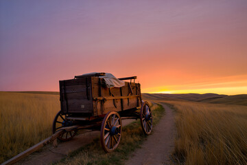 Fototapeta premium Old wooden wagon on a dirt path against a vibrant sunset over rolling prairie hills, evoking a sense of history and journey.
