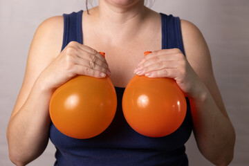 A woman holds two orange balloons in front of her chest, representing the effects of pregnancy and age on breasts. She examines the changes while reflecting on options for body image improvement.