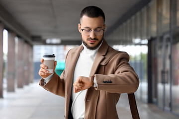 Young male businessman with coffee looking at wristwatch outdoors. Time management concept