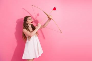 Charming woman in white dress holding a heart-shaped arrow with a playful gesture on a pink background