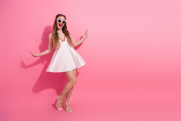 Young woman in stylish white dress and heart-shaped glasses posing against vibrant pink background...